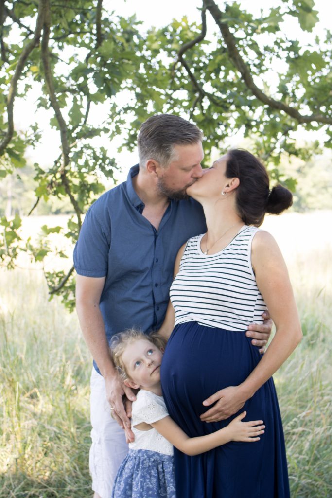Familienportrait mit einer schwangeren Frau, ihrem Mann und der Tochter unter einem Baum auf einer Wiese stehend.