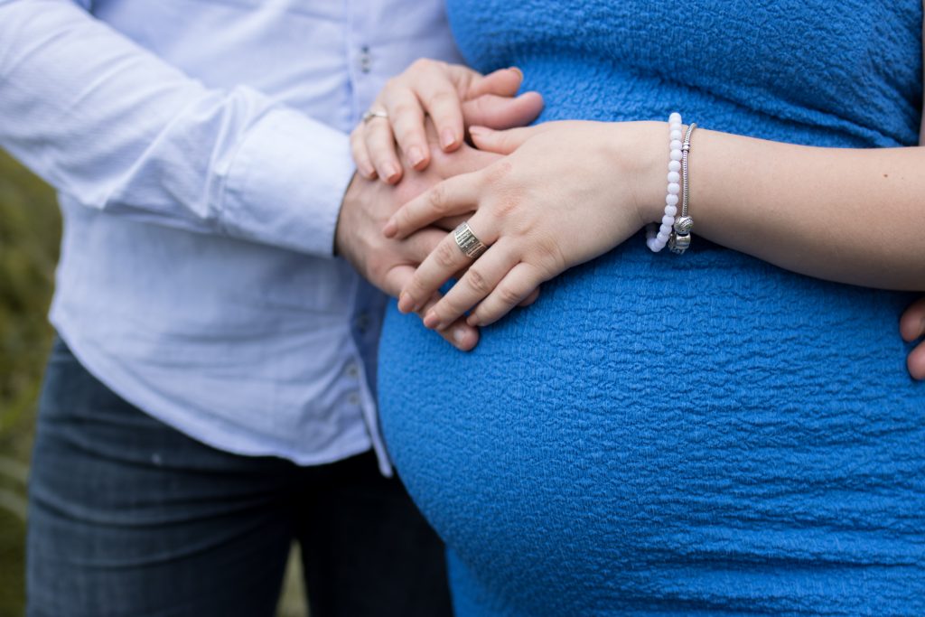 Paar hat beide Hände auf dem Babybauch der Frau, welche ien Blaues Kleid trägt.