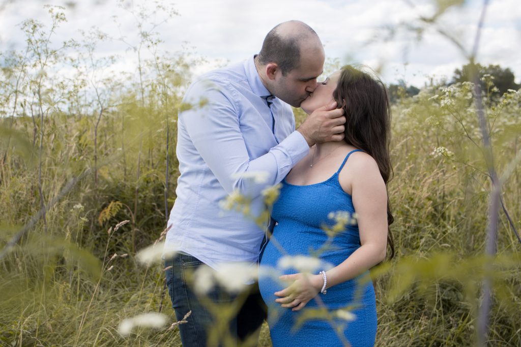 Paar küssend, Frau mit Babybauch an der Elbe in hoher Wiese stehend mit blauem Kleid.