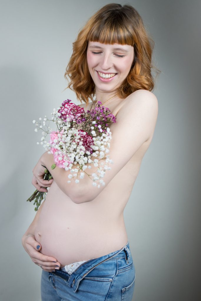 Schwangere Frau oben ohne KLeidung mit Blumen vor den Brüsten im Arm, in Jeans und streichelt den Bauch mit der anderen HAnd.