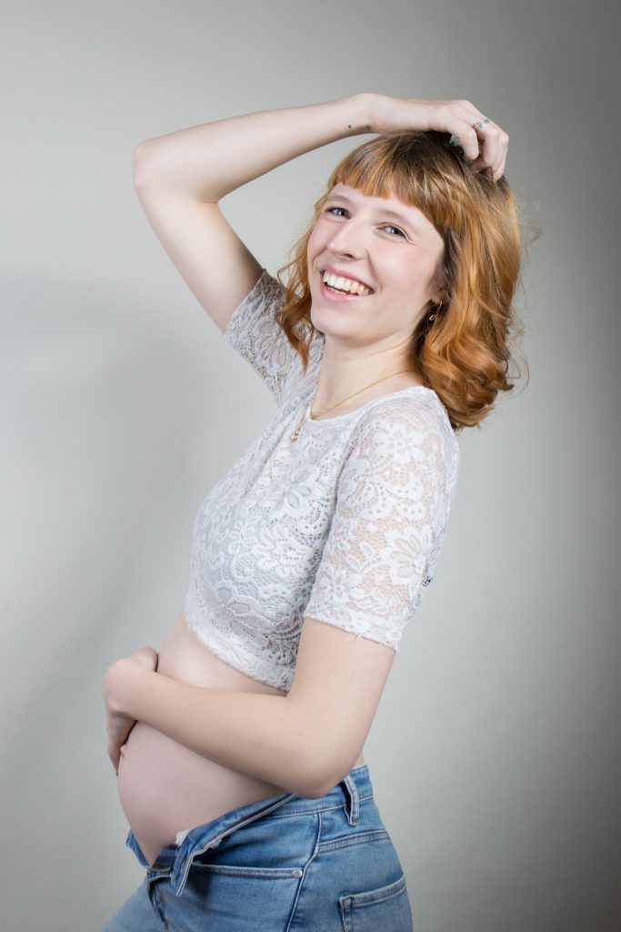Schwangere Frau mit Blumenmuster Croptop und Jeans vor weißem Hintergrund.