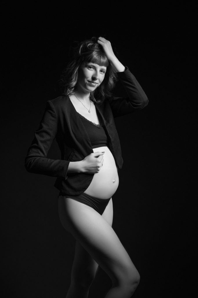 Schwangere Frau in schwarzer Unterwäsche mit einem schwarzen Blazer beim artistischen Schwangerschaftsfoto mit schwarzem Hintergrund