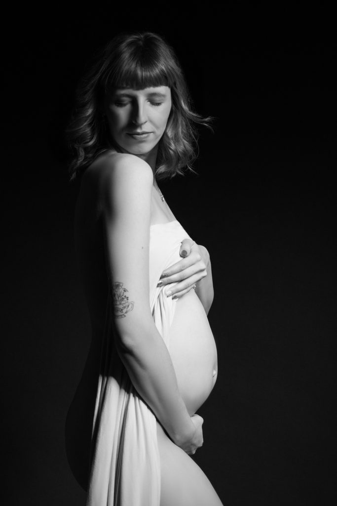Schwangere Frau in weißem Seidenkleid beim artistischen Schwangerschaftsfoto mit schwarzem Hintergrund