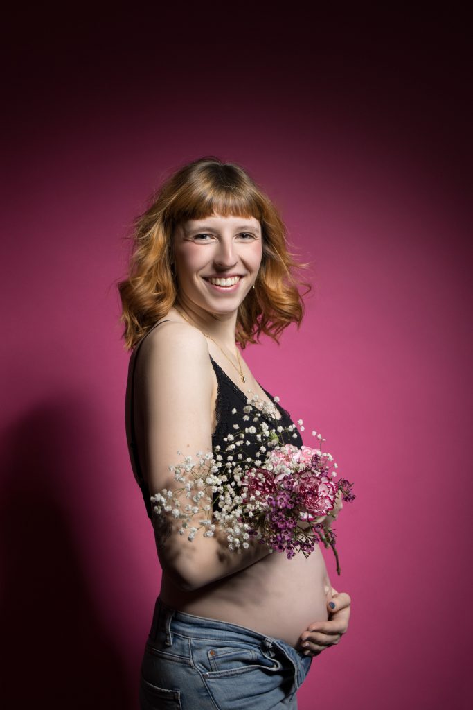 schwangere Frau in schwarzer Unterwäsche mit Blumen in den Armen vor Pinkem Hintergrund.