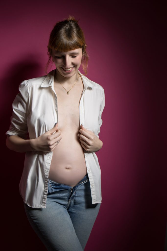 schwangere Frau ohen Unterwäsche mit einem weißem Hemd, was die Brüste bedeckt, und einer Jeans an vor Pinkem Hintergrund.