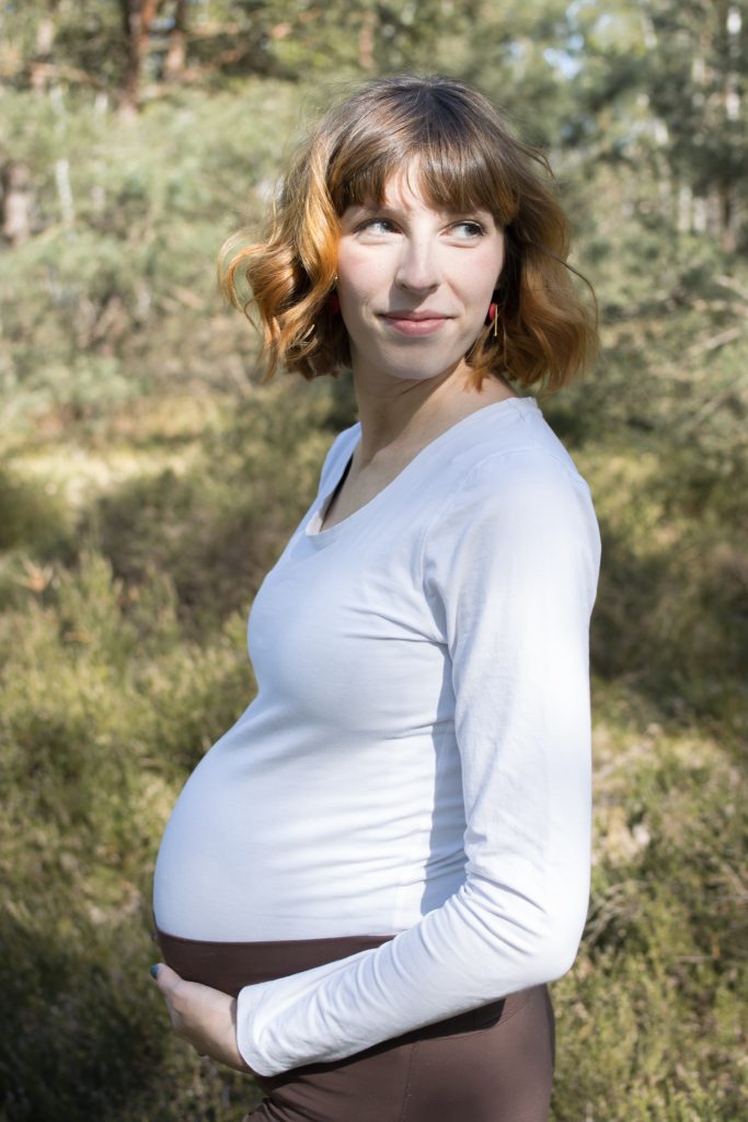 Eine schwangere Frau, welche sich den Babybauch hält, im neutralen Outfit im Wald auf einer Lichtung.